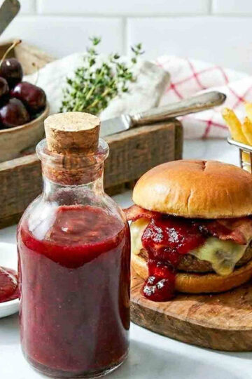 Bottle of homemade cherry ketchup served beside a bacon cheeseburger and fries, with fresh cherries and a small bowl of ketchup in the background.