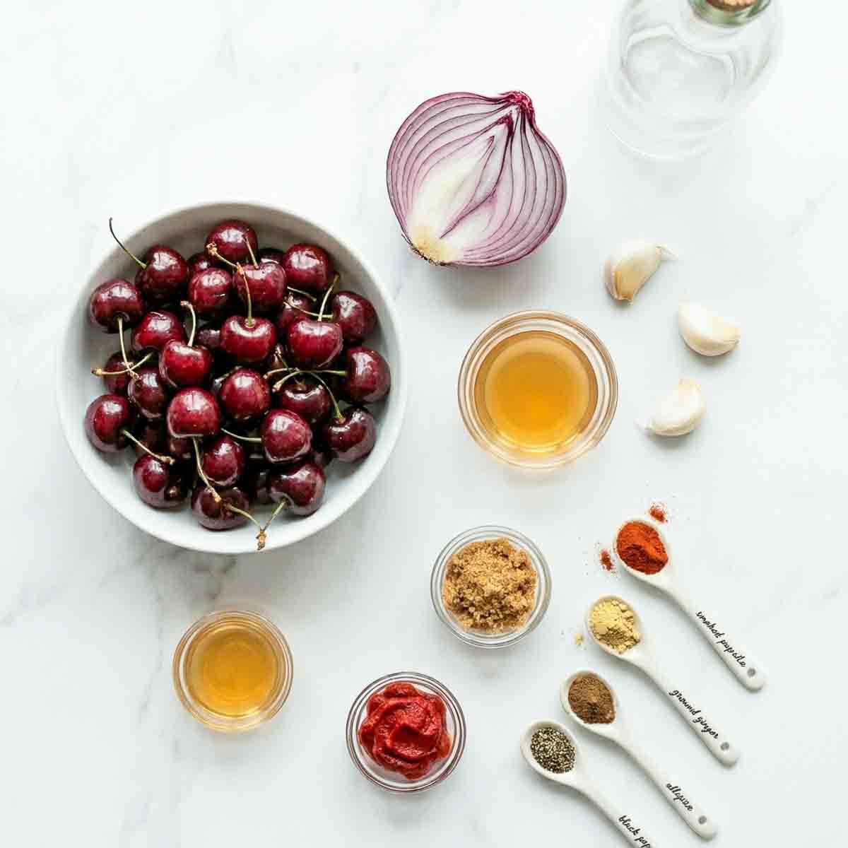 Ingredients for homemade cherry ketchup arranged on a marble surface, including fresh cherries, red onion, garlic, honey, apple cider vinegar, tomato paste, brown sugar, and spices.