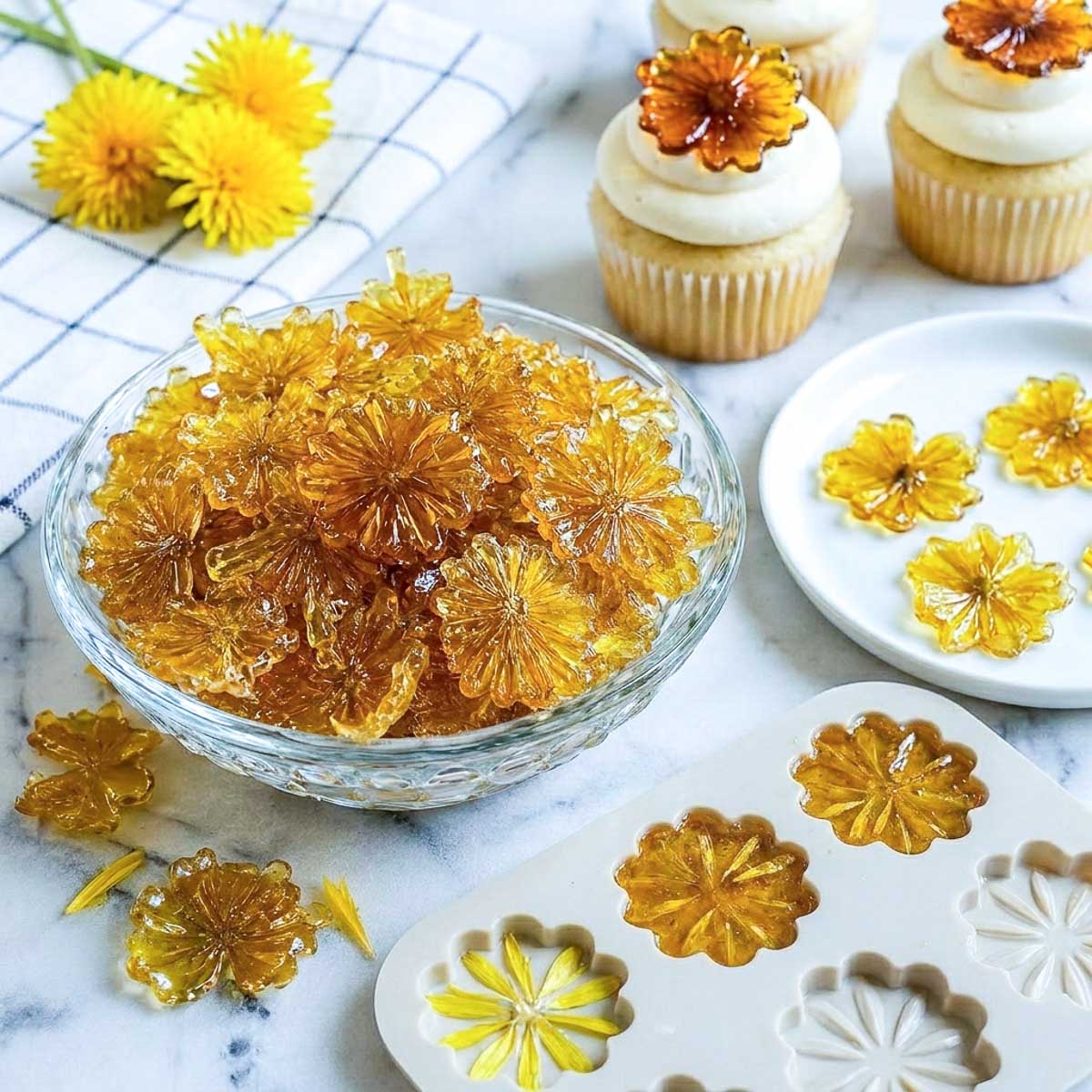 Homemade dandelion flower candy in amber-colored floral shapes arranged in a crystal bowl with cupcakes and silicone candy molds on a white marble surface.