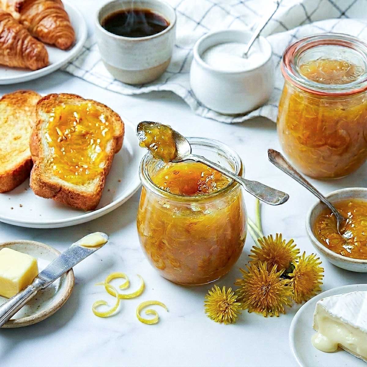 Jar of homemade dandelion flower jam with toasted bread, butter, croissants, and tea styled on a white marble background with fresh dandelion flowers.