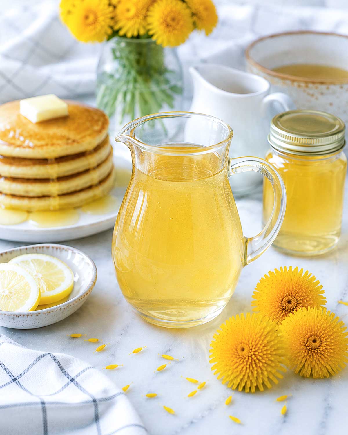 Glass pitcher of pale golden homemade dandelion syrup served beside pancakes, lemon slices, and fresh dandelion flowers on a white marble surface.