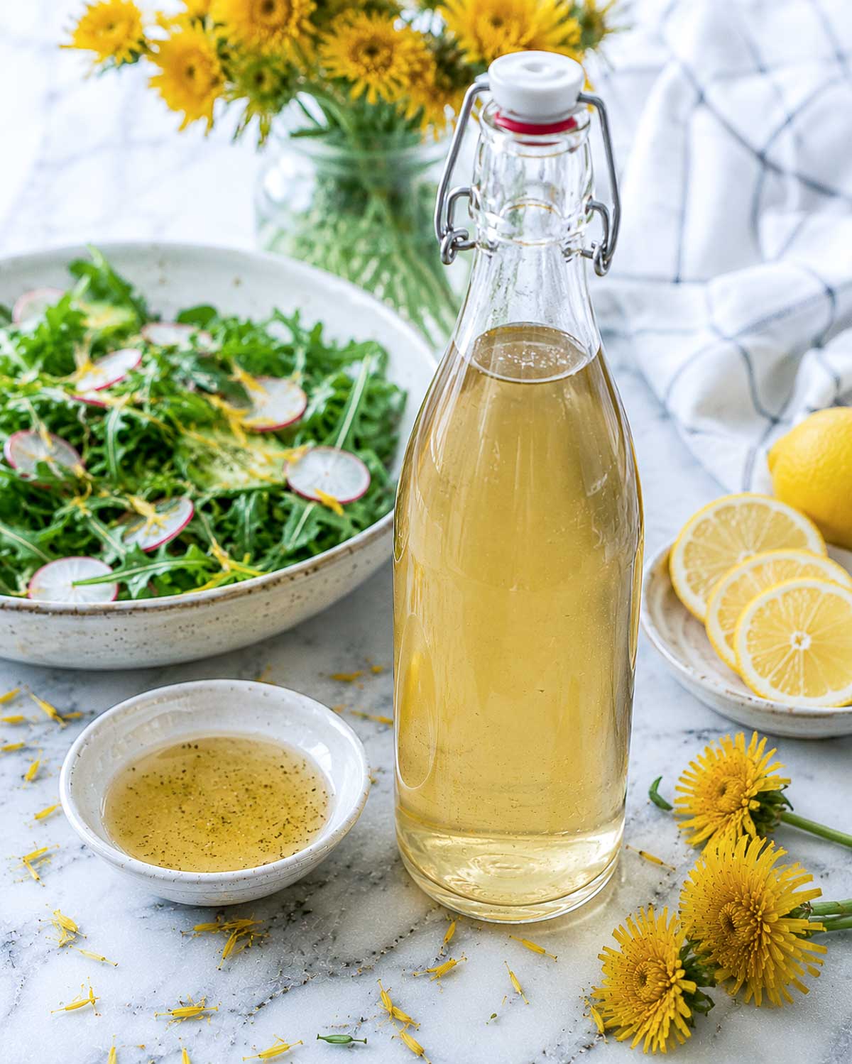 Bottle of homemade dandelion vinegar served with fresh salad, lemon slices, and dandelion flowers on a bright white marble background.