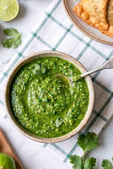Raw mango chutney recipe in a ceramic bowl with a silver spoon, samosas, lime halves, cumin seeds, cilantro, and green chilies on a marble surface.