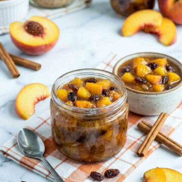 Glass jar of chunky peach chutney with raisins and cinnamon sticks on a white marble surface, styled with fresh peach slices and a soft orange check napkin.
