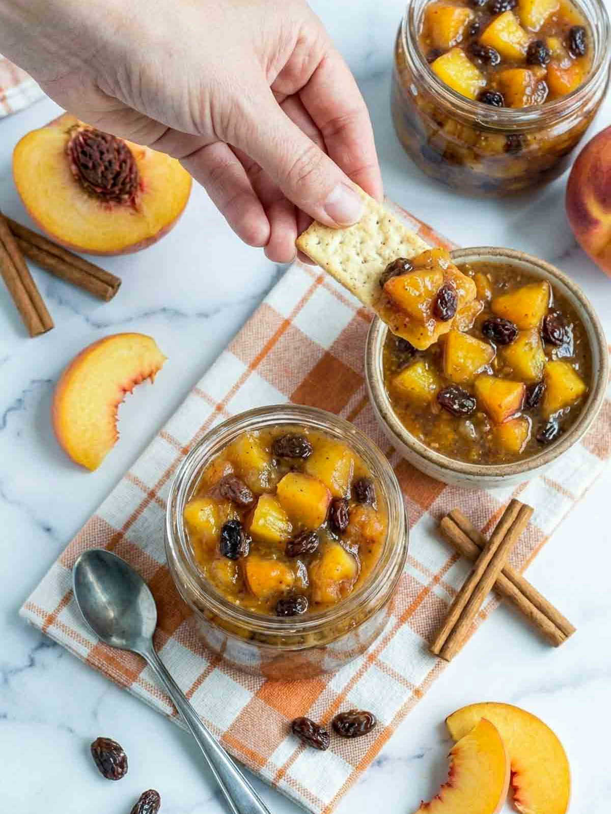 Hand dipping a cracker into chunky peach chutney with raisins and warm spices, styled with fresh peaches and cinnamon sticks on a marble surface.