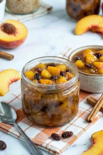 Close side view of homemade peach chutney in a glass jar with visible peach chunks and raisins, surrounded by cinnamon sticks and fresh peach slices.
