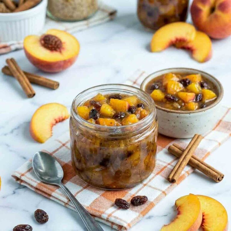 Close side view of homemade peach chutney in a glass jar with visible peach chunks and raisins, surrounded by cinnamon sticks and fresh peach slices.