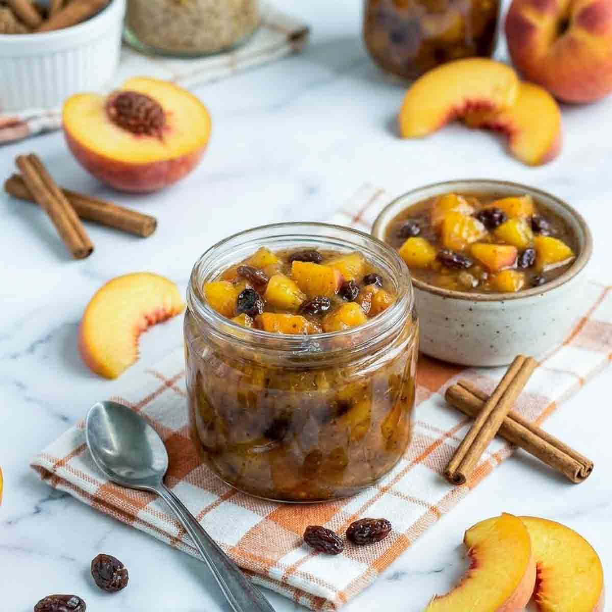 Close side view of homemade peach chutney in a glass jar with visible peach chunks and raisins, surrounded by cinnamon sticks and fresh peach slices.
