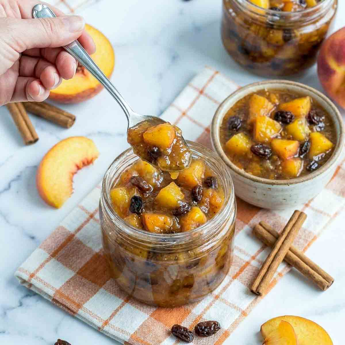 Spoon lifting thick peach chutney with raisins from a glass jar, styled with cinnamon sticks, fresh peaches, and a soft check napkin.